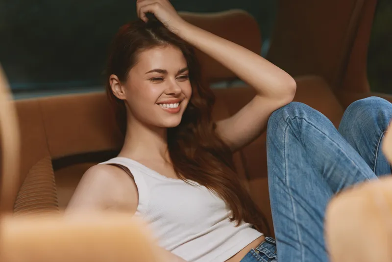 Joyful young woman smiling in casual outfit sitting in a car, showcasing a relaxed vibe with denim jeans and a tank top, perfect for travel or lifestyle themes.