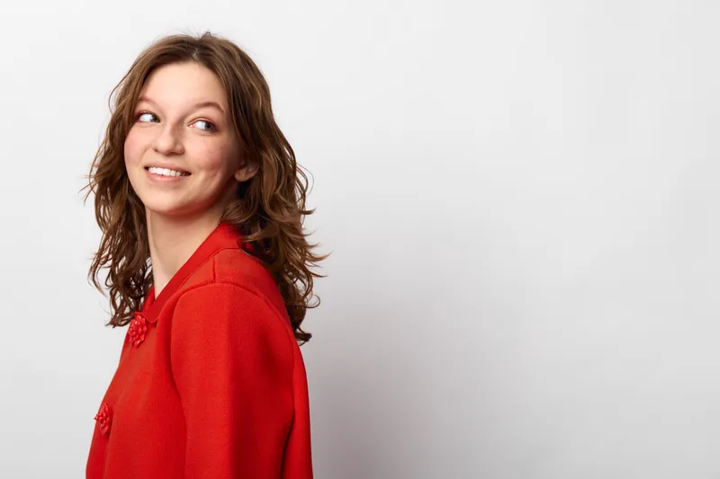 Young woman with curly brown hair wearing a red sweater looks sideways with a smile on a white background. Concept of casual style, happiness, and natural beauty.