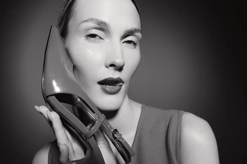 A fair skinned European woman with slicked back hair holds a high heel shoe near her face in a stylish black and white portrait. Concept of fashion and elegance.