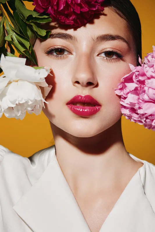 Young woman with glossy red lips poses with pink and white peonies near her face on yellow background. Clean skin and creative beauty concept with vibrant fashion style.