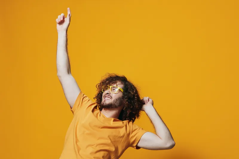 Happy young man with curly hair and yellow sunglasses dancing joyfully against a bright yellow background, showcasing vibrant energy and positivity