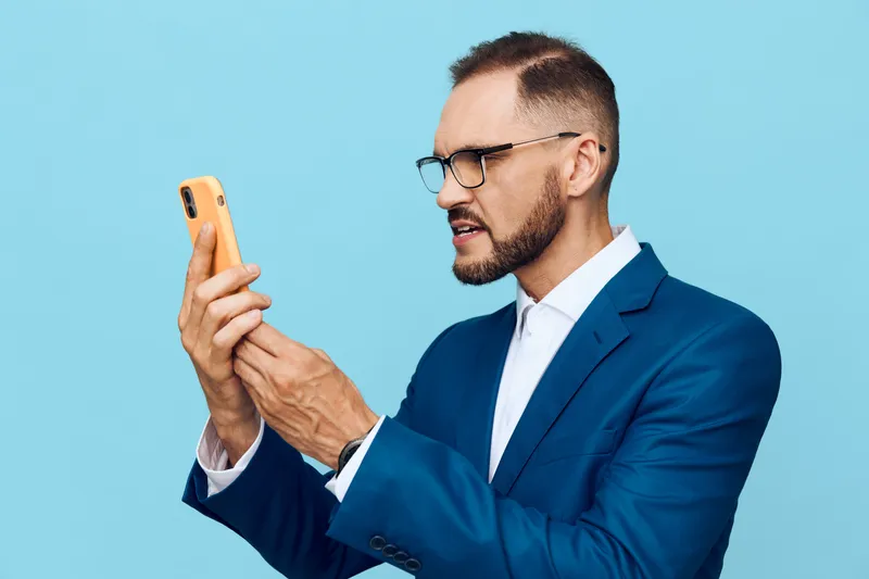 Professional businessman on a solid blue background, focused expression while checking a smartphone. This isolated color setup emphasizes ambition, concentration and confidence.