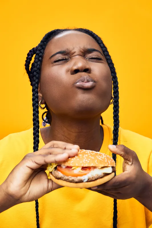 Happy young woman enjoying a delicious burger against a bright yellow background, showcasing her love for fast food with a smile and playful expression