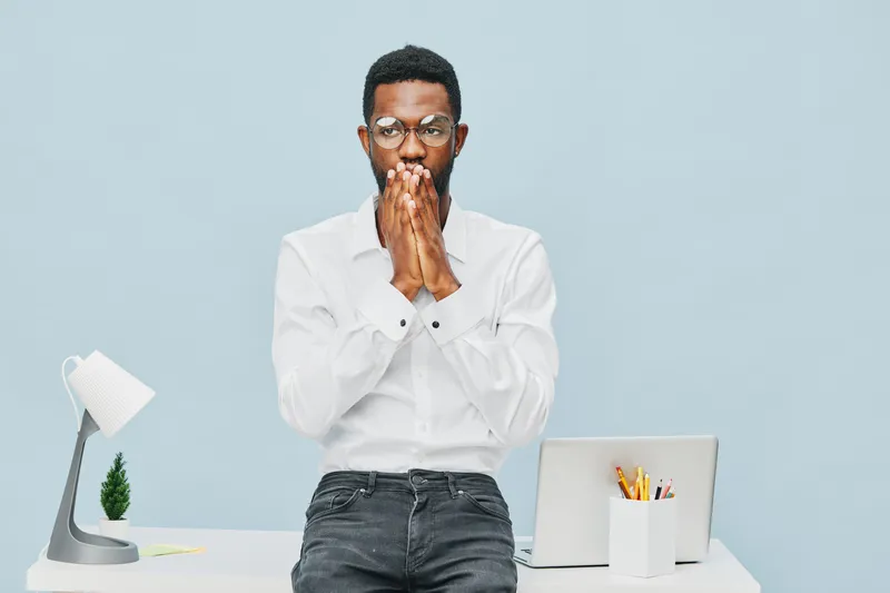 Confused young African American man in white shirt and glasses standing at modern white desk with laptop and stationery, touching his face with both hands in front of plain light blue background. Peop