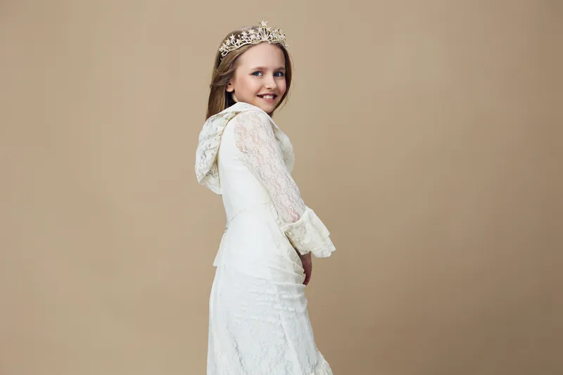Beautiful young girl in a lace dress with a crown, smiling joyfully against a beige background, capturing elegance and innocence in a charming portrait
