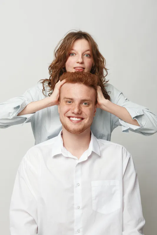 Smiling redhead man in white shirt sits while woman with curly hair playfully holds his head from behind. Studio portrait on light background with casual clothing and friendly mood.