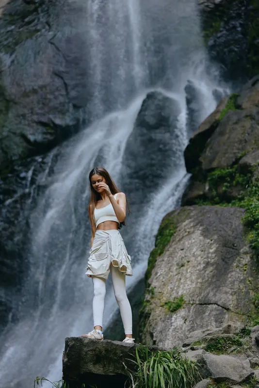 young woman standing near waterfall wearing white activewear and leggings in lush green nature, outdoor adventure and fitness lifestyle