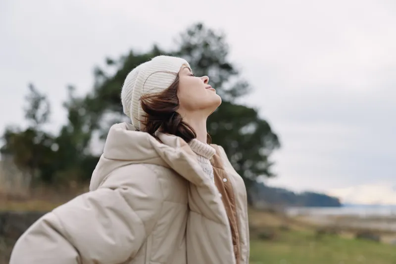 Outdoor portrait of a person in a beige coat and beanie, looking up at the sky with a calm expression, open landscape with trees and distant water under soft light.