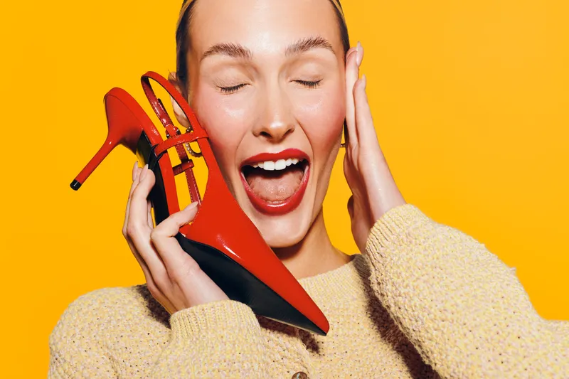 Happy woman with closed eyes holds red high heel shoe near face, smiling with open mouth on bright yellow background. Concept of fashion, style, and joyful emotions.