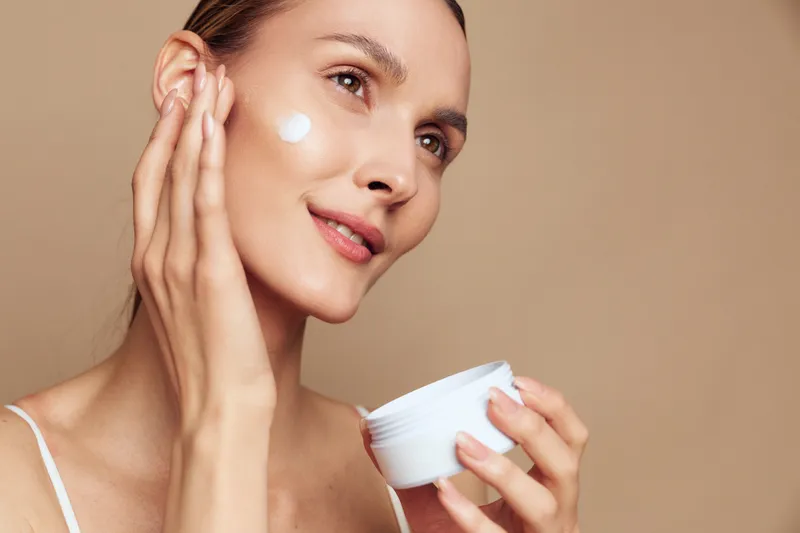 Young woman with clear skin applying white face cream on cheek, holding cream container. Beige background, natural makeup, concept of skincare, beauty, and self-care routine.