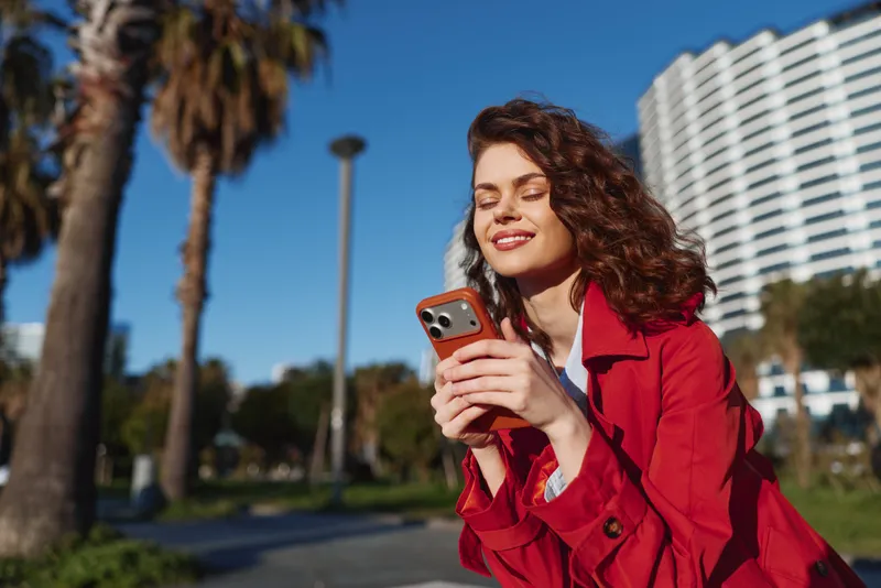 A young woman in a red coat smiles while scrolling on her smartphone in a sunny urban park, capturing a candid moment of connection and joy.