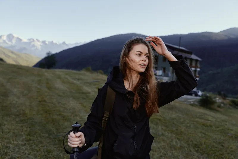 A calm outdoor hiker is seen trekking on a grassy hillside with mountains in the background. She looks focused and confident, creating an adventurous and tranquil atmosphere.
