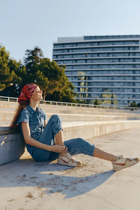 Lifestyle woman in modern boho-western denim street style relaxing outdoors with film color aesthetic, casual sneakers, and red bandana under bright sunlight.