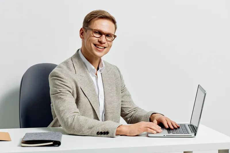 smiling young man working on laptop wearing glasses beige jacket seated at white desk in modern minimal office interior digital nomad remote work concept