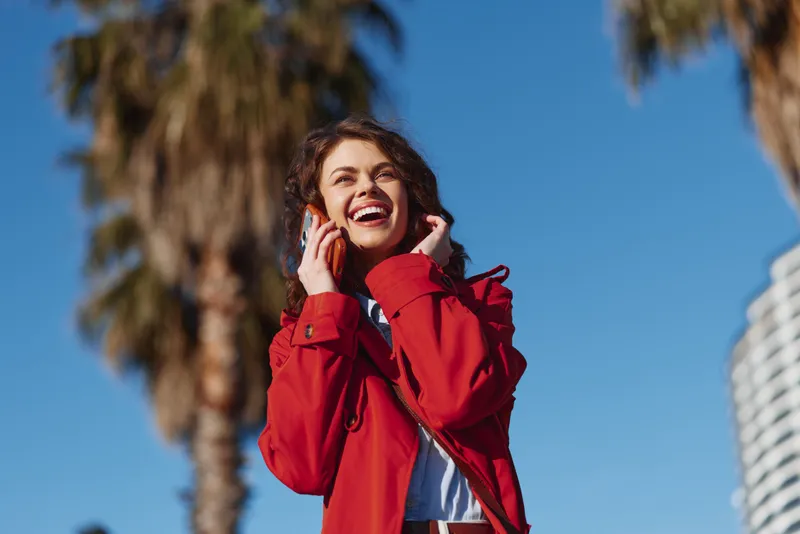 Young woman in a red coat laughs joyfully while talking on a smartphone outdoors on a sunny day with palm trees in the background.
