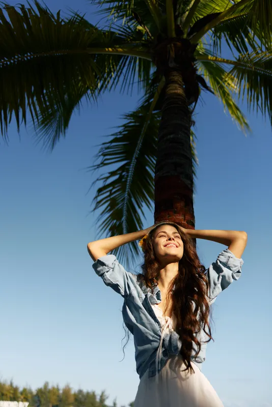 A woman stands beneath a tall palm tree, arms lifted to sunlit sky, wearing a casual denim jacket. This moment captures carefree summer vibes and warmth.