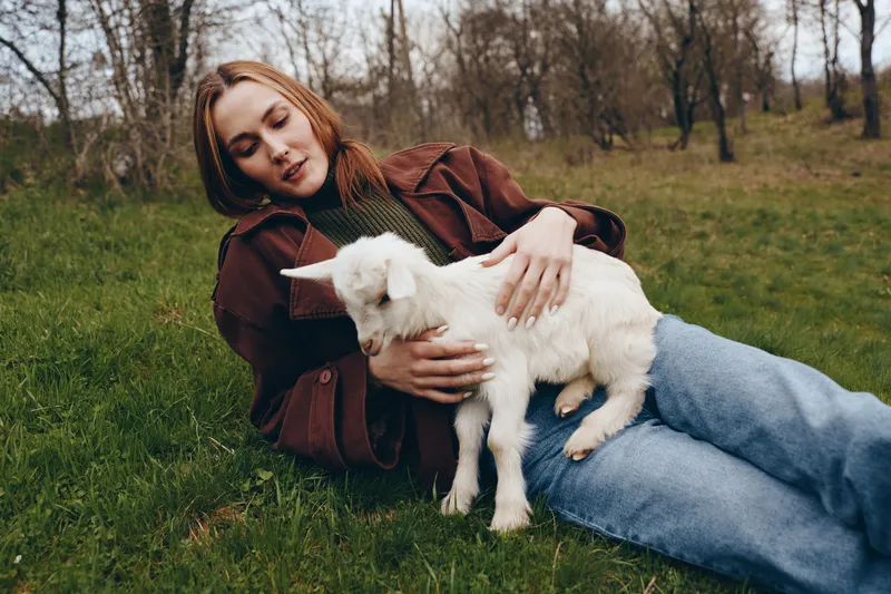 Calm woman lying on grass outdoors, cuddling a young goat kid, warm sunlit scene with natural tones, relaxed posture, soft breeze, intimate countryside vibe, rustic fashion