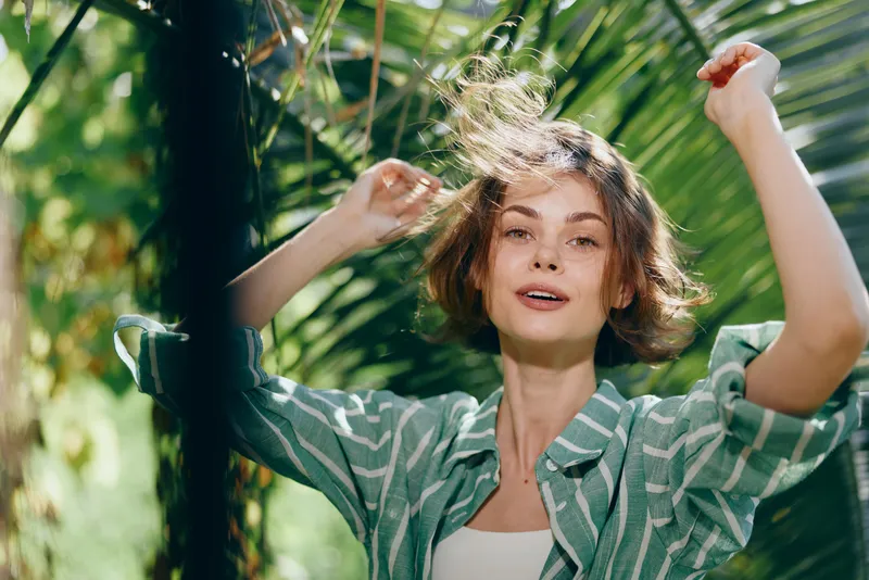 Joyful woman dancing outdoors among lush tropical plants, sunlight glints on her hair, wearing a casual striped shirt, conveying carefree energy and vibrant summer mood.