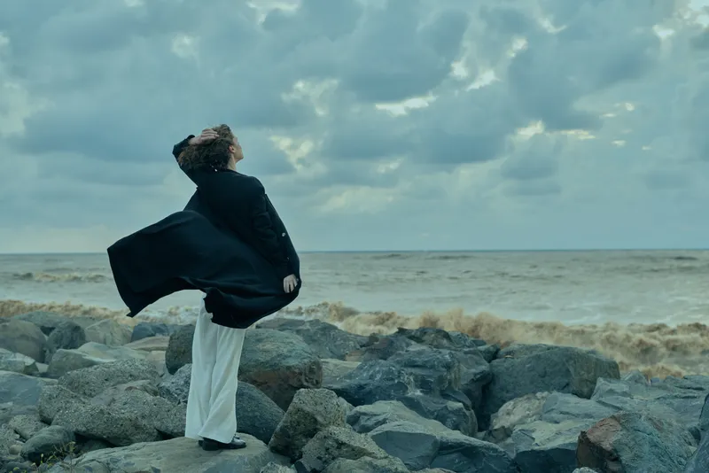Wind swept scene with cloak on rocky shore, waves in the sea behind, solitary figure facing the horizon in dramatic coastal landscape