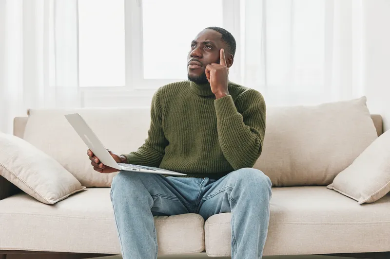 Thoughtful black man in green sweater sitting on sofa with laptop, contemplating ideas in a bright room, showcasing creativity and inspiration