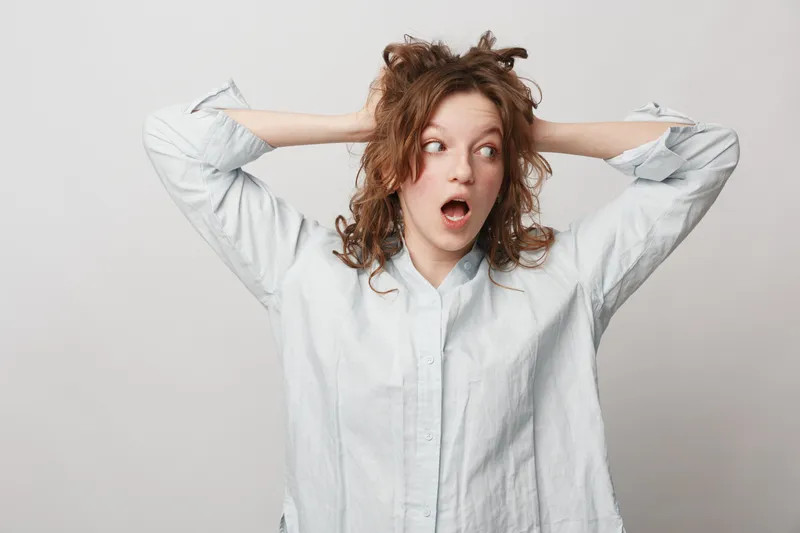 Surprised young woman with curly hair wearing white shirt holds hands behind head, looking to side with open mouth. Concept of shock, amazement, and unexpected reaction in casual style.