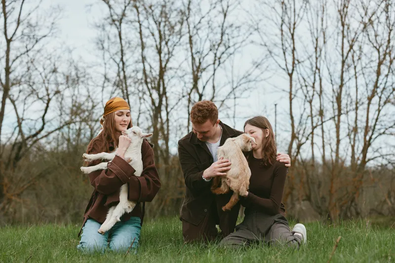 Smiling family outdoors with a goat kid, adults holding two young animals, in a green field with bare trees and warm daylight, portraying lifestyle, animals, togetherness, playful bonding and a