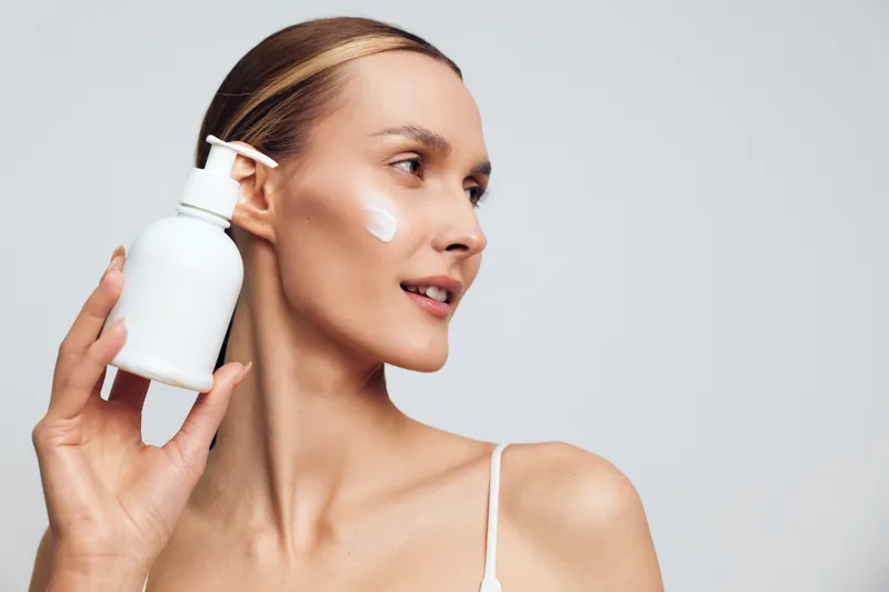 Young woman applying cream on face holding white lotion bottle skincare beauty routine clean fresh skin natural look minimal background