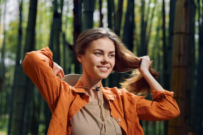 Woman smile forest hair outdoor nature portrait happy young woman adjusting her hair in an orange jacket with backpack, natural sunlight through bamboo trees and a relaxed, cheerful expression.