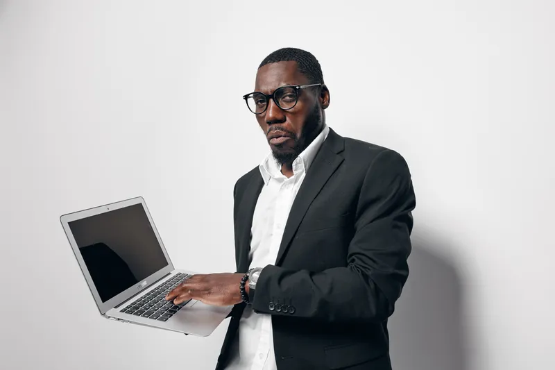 African American man with dark skin wearing glasses and black suit holding laptop computer standing against white background for business or technology concept.