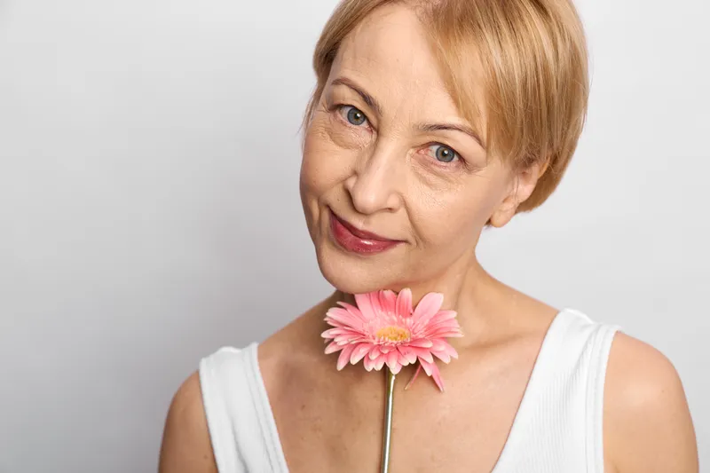 Senior woman with blond hair holding a pink flower close to her lips, gentle smile, studio portrait, soft lighting, clean white background, calm friendly expression, beauty mood
