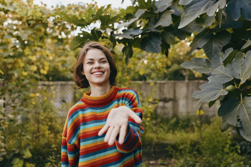 Woman smiling and reaching hand for handshake in colorful striped sweater outdoors in garden, portrait of invitation and connection, young person among green leaves and warm natural light.