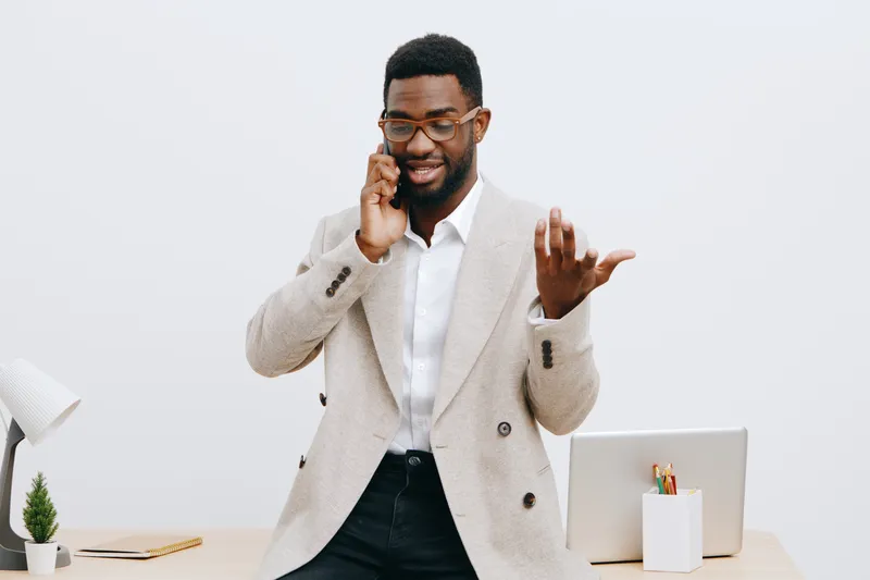 Stylish young African American man wearing beige blazer and white shirt, talking on mobile phone while smiling confidently in modern office interior, lifestyle business professional concept