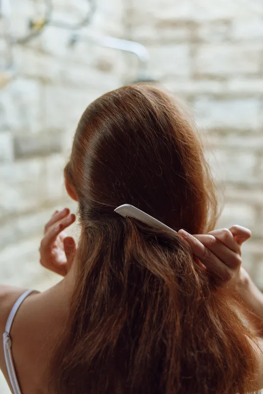Woman combing long brown hair in natural light, close-up shot with soft background, indoor setting, casual moment of self-care and grooming.