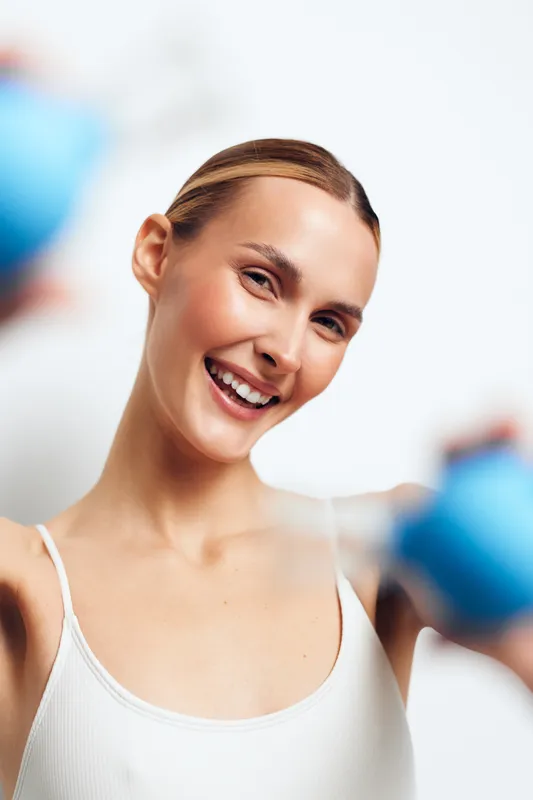 Smiling young woman in white tank top holding blue dumbbells exercising. Bright studio lighting highlights fitness, health, and active lifestyle concepts. Clean white background.