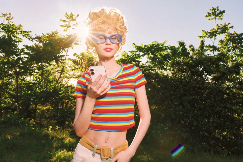 Young woman with curly blonde hair and blue sunglasses wears a colorful striped crop top in a sunny park while looking at her smartphone exuding playful retro vibes