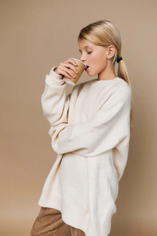 Softly dressed in an oversized cream sweater, a young woman drinks from a paper cup, capturing a calm moment of comfort and a quiet, cozy morning routine.