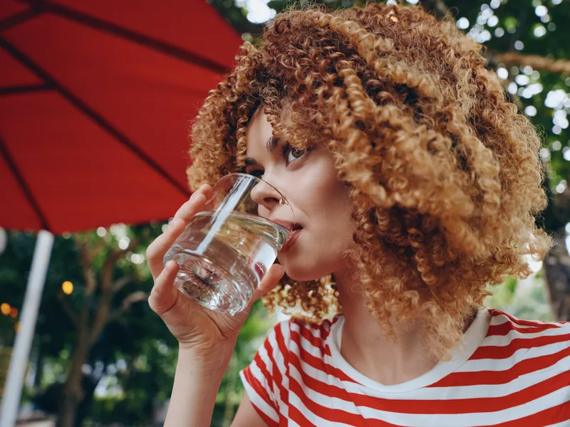 Portrait of a woman with curly hair drinking water from a glass outdoors, under a bold red umbrella, highlighting hydration, calm focus, and a casual summer moment.
