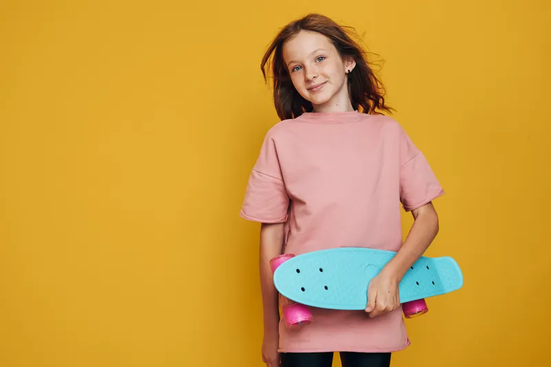 Smiling young girl in a pink shirt holding a blue skateboard standing against a yellow background. Concept for youth, sports, and casual lifestyle.
