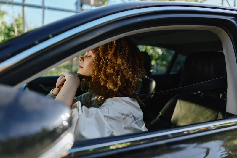 Woman with curly hair relaxing in black car, wearing casual white shirt, enjoying peaceful moment with closed eyes and natural light inside vehicle.