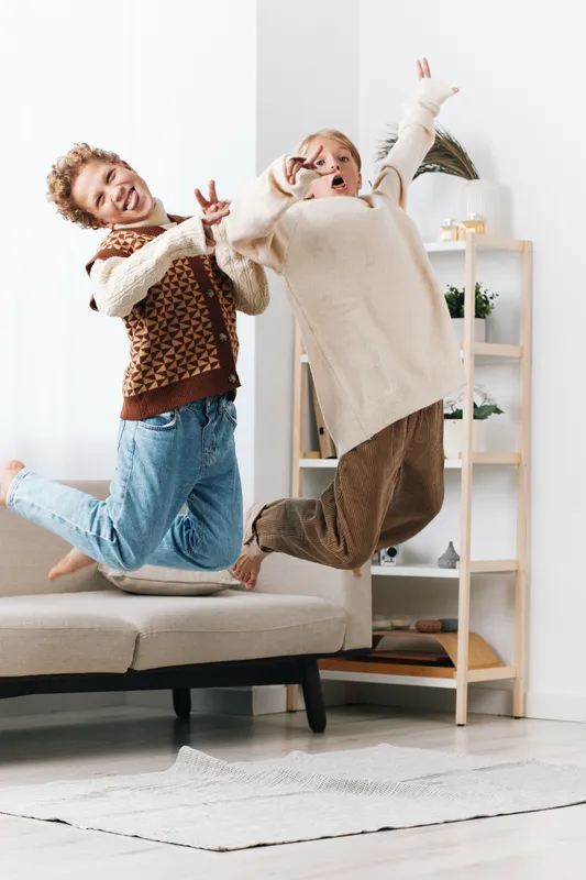 Authentic moment of playful motion as two friends jump on a sofa, reflecting a school age lifestyle and carefree play, capturing spontaneous joy in a bright room and soft light.