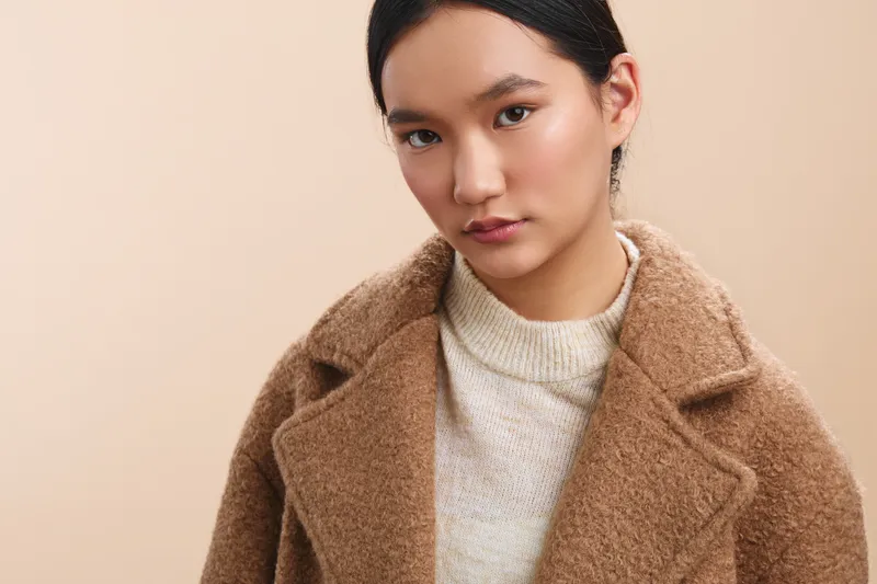 Asian woman wears a cozy coat and soft sweater, facing the camera with a neutral gaze. The clean studio setting highlights modern fashion sensibility and a calm, confident presence.