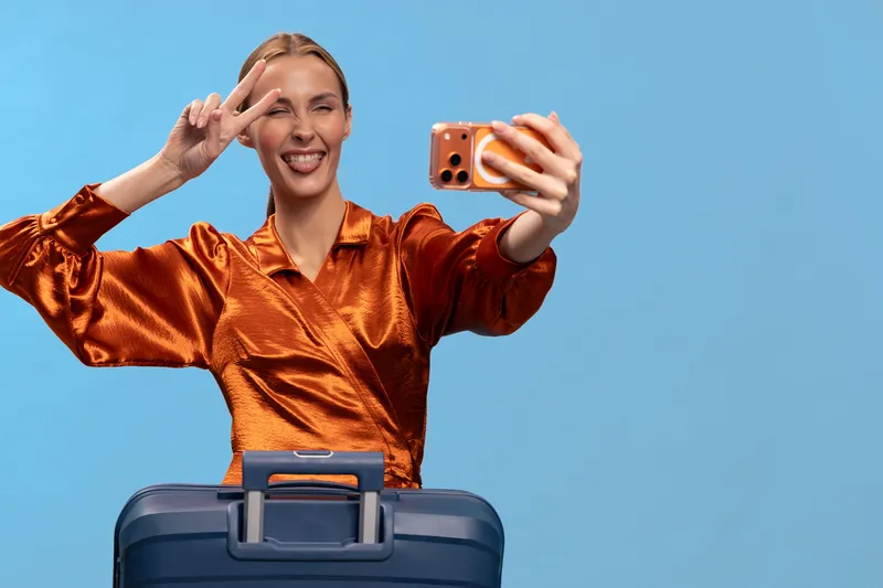 Young woman in shiny orange blouse takes selfie with smartphone and makes peace sign while standing behind blue suitcase on blue background. Concept of travel, vacation, and joyful mood.