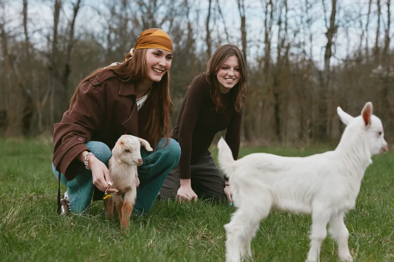 Two girls share a playful moment with a kid goat outdoors in the countryside, capturing a lifestyle scene with friends, animals and nature on a grassy field, bright smiles and candid interaction
