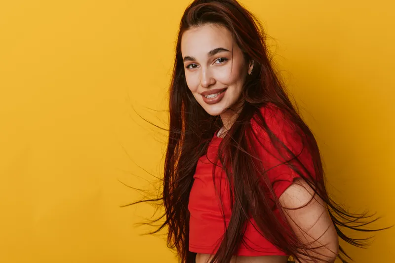 Young woman with long hair smiling and wearing red shirt standing against yellow background. Portrait of happy female with natural makeup and casual style in studio lighting.