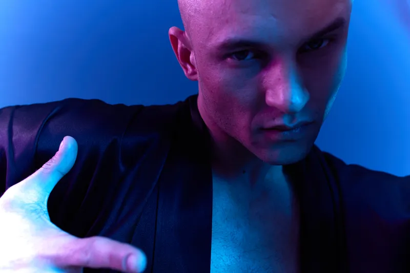 Confident man in a dark robe poses dramatically, showcasing strong emotions with vibrant blue and purple colors. Unique and striking portrait in modern style.