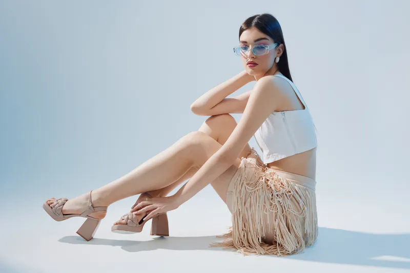 Young woman in stylish fashion outfit with fringe skirt and high heels poses sitting on floor against light background. Concept of beauty, fashion, and modern style in studio.
