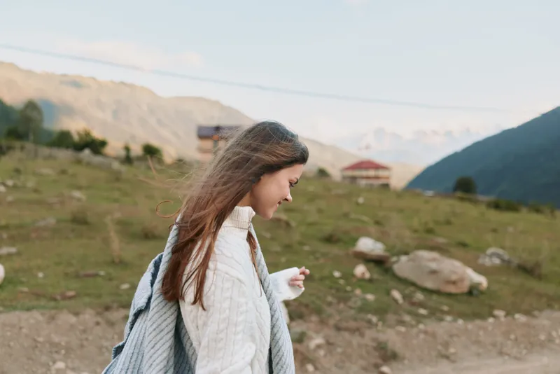 Young woman walks along a mountain trail in a cozy sweater, enjoying a calm outdoor moment amid rugged scenery and clear skies.