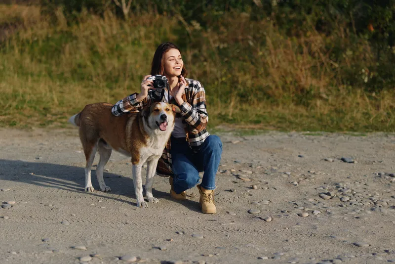 Woman, dog, photography, outdoors, camera, joyful moment on a pebble beach with a dog, smiling woman with camera, sunlight, nature backdrop, warm companionship