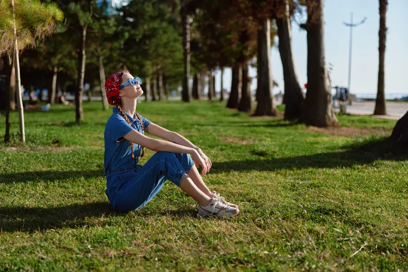 Lifestyle woman in modern boho-western denim street style sitting on grass outdoors, wearing sunglasses and a red bandana, enjoying sunny weather with film color effect.
