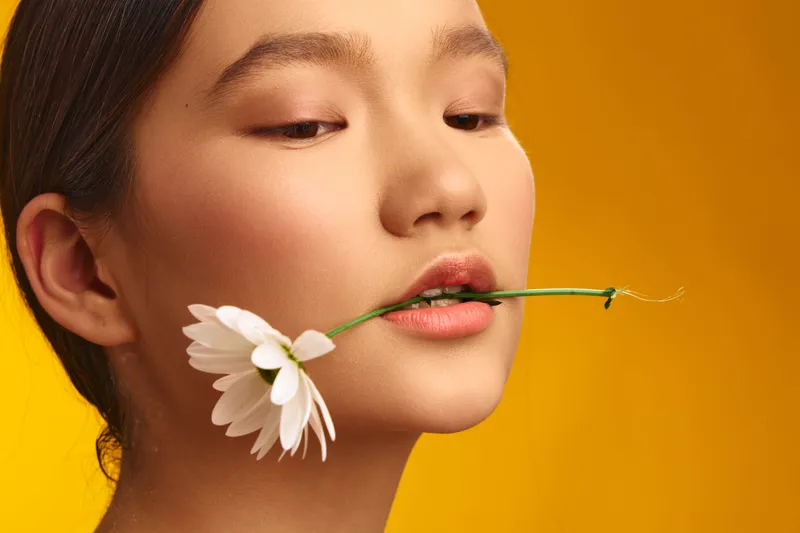 asian woman with a delicate daisy held between her lips, softly glowing studio light highlights smooth skin and poised features, creating a serene, fresh beauty with warm, inviting tones.